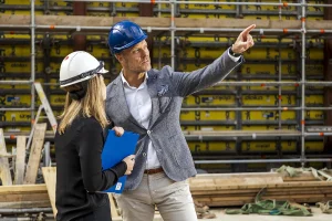 Een man en een vrouw, beiden met beschermende helmen, staan op een bouwplaats. De man, met een blauwe helm en een grijze blazer, wijst terwijl hij met de vrouw praat, die een blauwe map vasthoudt. Op de achtergrond zijn steigers en bouwmaterialen zichtbaar: een perfect decor voor bedrijfsfotografie.