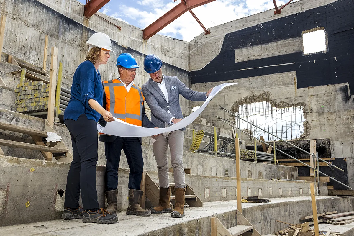 Drie mensen op een bouwplaats onderzoeken architectonische plannen. Eén persoon, met een witte helm, een blauw shirt en een spijkerbroek, staat naast twee anderen: een met een blauwe helm, een oranje vest en een grijze outfit; de ander met een blauwe helm, grijze blazer en kaki broek. Het gedeeltelijk gebouwde gebouw toont zijn betonstructuur. Ideaal voor bedrijfsfotografie!