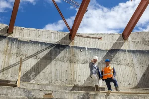 Twee bouwvakkers inspecteren een grote bouwplaats. Beiden dragen een veiligheidshelm, de een draagt een grijs pak en de ander draagt een oranje veiligheidsvest. Stalen balken hangen boven het hoofd en onafgewerkte betonnen muren omringen ze. De blauwe lucht met wolken vormt een ideaal decor voor bedrijfsfotografie.