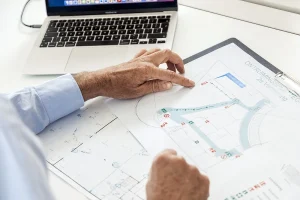 Een persoon in een blauw shirt wijst met één hand naar een blauwdruk op een klembord, terwijl de andere hand vlakbij rust. De blauwdruk bevat diagrammen en symbolen. Op het bureau boven de blauwdruk ligt een laptop met een gedeeltelijk zichtbaar scherm open, een ideaal moment voor bedrijfsfotografie.