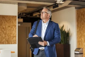 Een man van middelbare leeftijd met kort grijs haar en een bril, gekleed in een blauwe blazer en een wit overhemd, houdt een zwarte portefeuille en pen vast terwijl hij nadenkend naar boven kijkt. Hij staat in een moderne kantoorruimte met houten en witte muren, een ideale setting voor bedrijfsfotografie, met een grote potplant op de achtergrond.