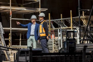 Twee mannen met veiligheidshelmen staan op een bouwplaats. Eén, gekleed in een blauwe blazer en een lichte broek, wijst in de verte. De ander, in veiligheidsvest en donkere kleding, kijkt dezelfde kant op. Op de achtergrond zijn steigers, metalen staven en bouwmachines zichtbaar: perfect voor bedrijfsfotografie.