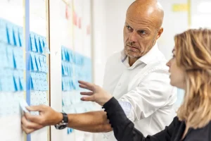 Een kale man in een wit overhemd en een vrouw met schouderlang haar raken in gesprek, wijzend naar kleurrijke plakbriefjes op een whiteboard. Het whiteboard is gevuld met blauwe sticky notes, gerangschikt in rijen, die de suggestie wekken van een gezamenlijke brainstormsessie of een projectplanningssessie: een ideale scène voor bedrijfsfotografie.