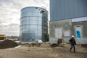 Een bouwplaats met een hoge, cilindrische metalen opslagtank naast een groot, gedeeltelijk gebouwd gebouw. Op de voorgrond loopt een arbeider met een harde hoed en een veiligheidsvest. De grond is oneffen met stukken vuil. Op de achtergrond staat een geel bouwvoertuig geparkeerd, dat door middel van bedrijfsfotografie de industriële vooruitgang laat zien.