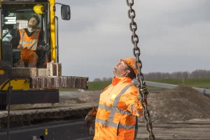 Twee bouwvakkers in oranje veiligheidsjassen en helmen zijn ter plaatse. De één bedient een gele hydraulische graafmachine, terwijl de ander naar boven kijkt en een zware ketting vasthoudt. De achtergrond bestaat uit een zandheuvel en een gedeeltelijk bewolkte hemel, perfect voor dynamische bedrijfsfotografie.