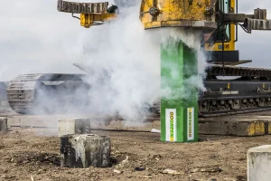 Een grote geel-zwarte machine drukt op een bouwplaats een groene stapel met het opschrift "GoGreen" in de grond. Rook of stoom omhult de machinerie en legt een industriële essentie vast, perfect voor bedrijfsfotografie. De onverharde grond is bezaaid met betonblokken, terwijl onder een bewolkte hemel een rupsband zichtbaar is.