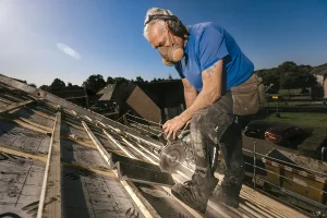 Een bouwvakker met grijs haar, gekleed in een blauw shirt, masker, gehoorbescherming en gereedschapsriem, zaagt met een cirkelzaag houten planken op een schuin dak onder een helderblauwe lucht. Op de achtergrond zijn huizen en bomen zichtbaar. Rondom de zaag is stof en zaagsel te zien in dit voorbeeld van bedrijfsfotografie.