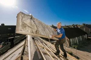 Een arbeider in een blauw shirt en een donkere broek installeert op een zonnige dag grote isolatiepanelen op een schuin dak van een woongebouw. De arbeider, vastgelegd met nauwkeurige bedrijfsfotografie, staat aan de rand van het dak en tilt het isolatiemateriaal op. Op de achtergrond zijn meerdere huizen en een straat zichtbaar.