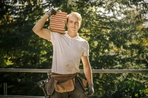 Buiten op een bouwplaats staat een man met een stapel stenen op zijn schouder. Hij draagt een wit poloshirt, een leren gereedschapsriem, handschoenen en een donkere broek. De achtergrond toont een groen, zonovergoten bos met metalen balustrades achter hem zichtbaar. Dit tafereel geeft perfect de essentie van bedrijfsfotografie weer.