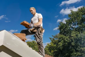 Een bouwvakker in een wit overhemd, een grijze broek met zakken en handschoenen staat op een dak met een stapel kleitegels in zijn hand. Om hun middel zit een gereedschapsriem. De lucht is helder met verspreide wolken en weelderige groene bomen omlijsten de achtergrond: een perfecte scène voor bedrijfsfotografie.