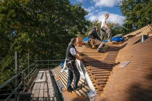 Drie bouwvakkers repareren op een zonnige dag een dak. Twee knielen op het pannendak en leggen nieuwe pannen, terwijl één op een nabijgelegen dakladder staat. Op de achtergrond zijn bomen en een blauwe lucht met wat wolken te zien. Steigers rondom het huis vormen een perfect moment voor bedrijfsfotografie.