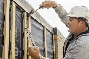 Op een bouwplaats wordt gezien hoe een man met een witte veiligheidshelm en een grijze capuchon een spijker in houten planken slaat. Met een waterpasinstrument in zijn andere hand werkt hij met precisie. De planken zijn bevestigd aan een bouwmuur met daarachter een zwarte ommanteling. De bewolkte lucht vormt het decor voor dit tafereel van bedrijfsfotografie.