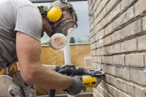 Een bouwvakker, vastgelegd in bedrijfsfotografie, draagt een grijs shirt, gele oorkappen, oogbescherming en een stofmasker terwijl hij geel elektrisch gereedschap gebruikt op een bakstenen muur. Handschoenen beschermen hun handen. Op de achtergrond zijn steigers en een bouwconstructie zichtbaar. De persoon is gefocust op zijn taak.