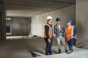 Drie mannen in bouwuitrusting staan op een bouwplaats plannen te bespreken. Twee dragen oranje veiligheidsvesten, witte overhemden en blauwe helmen; men leunt tegen een richel. De derde draagt een grijze jas, een blauw overhemd en een witte helm. Het bouwwerk, vastgelegd via bedrijfsfotografie, is onafgewerkt met betonnen muren en verspreide materialen.