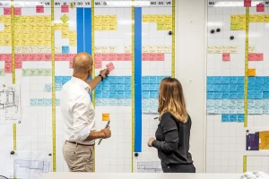 Twee mensen staan voor een whiteboard waarop verschillende gekleurde sticky notes in kolommen staan. De persoon links, kaal en gekleed in een wit overhemd, wijst naar een roze briefje, terwijl de persoon rechts, met lang bruin haar, toekijkt. Deze scène legt effectieve bedrijfsfotografie vast met aantekeningen geordend op dagen van de week.