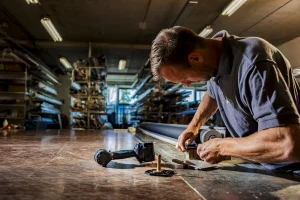 Een man met kort haar en baard werkt aandachtig aan een grote houten tafel in een goed verlichte werkplaats. Diverse gereedschappen en materialen, waaronder een boormachine, liggen verspreid op de tafel. Op de achtergrond zijn planken gevuld met lange cilindervormige voorwerpen en andere benodigdheden zichtbaar, die de essentie van bedrijfsfotografie vastleggen.