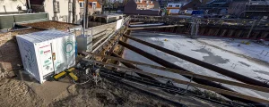 Bouwterrein met zware balken die een uitgravingsgebied ondersteunen. Op de voorgrond staat een gelabelde container met kabels en connectoren. De grond is vuil met een paar verspreide gereedschappen. Onder een heldere hemel zijn gebouwen en woonhuizen op de achtergrond zichtbaar, perfect voor bedrijfsfotografie.