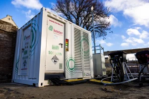 Buiten staat een grote witte container met het opschrift "kenter" en "500 kWh". Er zijn meerdere ventilatieopeningen, bedieningspanelen en kabels op de container aangesloten. Het staat op een omheind terrein met bouwmaterialen in de buurt, onder een helderblauwe lucht met wat wolken. Bomen en gebouwen staan op de achtergrond, perfect voor bedrijfsfotografie.