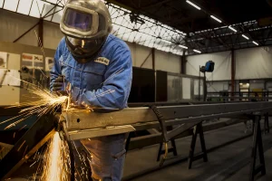 Een arbeider in een blauw beschermend pak en helm gebruikt elektrisch gereedschap om een grote metalen balk te slijpen, waardoor vonken ontstaan. De werkruimte, vastgelegd in prachtige bedrijfsfotografie, is een industriële omgeving met metalen constructies en apparatuur, verlicht door natuurlijk licht van grote ramen en plafondlampen.