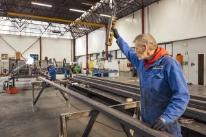 In een fabrieksomgeving, vastgelegd via bedrijfsfotografie, bedient een man, gekleed in een blauwe jumpsuit met rode accenten en zwarte handschoenen, een zwaar stuk gereedschap op een lange metalen balk. Op de achtergrond zijn andere werknemers zichtbaar, die verschillende taken uitvoeren onder hoge plafonds en grote ramen vol apparatuur en machines.