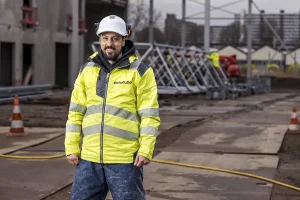 Een man in een felgeel veiligheidsvest en witte veiligheidshelm staat op een bouwplaats. Achter hem staan oranje verkeerskegels, metalen constructies en verschillende andere arbeiders in soortgelijke goed zichtbare kledij. De grond is oneffen, met overal verspreide kabels en bouwmaterialen: perfect voor bedrijfsfotografie.