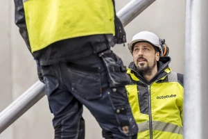 Twee bouwvakkers werken samen op een werkplek. De werker op de voorgrond, met zijn gezicht naar de andere kant gericht, draagt een zwarte broek en een geel goed zichtbaar jack. De andere arbeider, die een soortgelijke jas en een witte helm draagt, kijkt naar hem op. Ze zijn omgeven door metalen steigers, waardoor een authentiek moment in bedrijfsfotografie wordt vastgelegd.