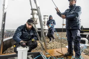 Drie werknemers op een met grind bedekt dak installeren of onderhouden apparatuur onder een bewolkte hemel. Eén knielt en hanteert een cilindervormig apparaat, terwijl de andere twee kabels en kettingen beheren. Ze dragen beschermende kleding en handschoenen, wat de precisie weerspiegelt die vaak in bedrijfsfotografie wordt vastgelegd.