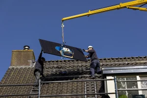 Twee arbeiders installeren zonnepanelen op een pannendak onder een helderblauwe hemel. Een gele kraan ondersteunt een van de panelen in de lucht, terwijl een medewerker op de steiger het bestuurt en de ander zich voorbereidt om het vast te zetten. Beide werknemers dragen veiligheidsuitrusting, inclusief helmen en harnassen: perfect voor bedrijfsfotografie.