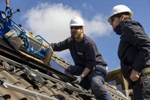 Twee arbeiders met helmen en beschermende uitrusting installeren zonnepanelen op een pannendak onder een gedeeltelijk bewolkte hemel. Eén arbeider knielt en bedient de installatieapparatuur, terwijl de ander vlakbij staat te observeren. De scène belichaamt hernieuwbare energie en vangt de essentie van bedrijfsfotografie.