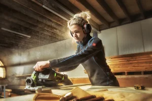 Een persoon met blond haar dat naar achteren is getrokken, oorbeschermers en een zwarte jas draagt, bedient een groene elektrische zaag in een houtwerkplaats. Zaagsel hangt in de lucht en op de achtergrond zijn houten planken zichtbaar. Op de voorgrond liggen verschillende handgereedschappen uitgespreid op de tafel.