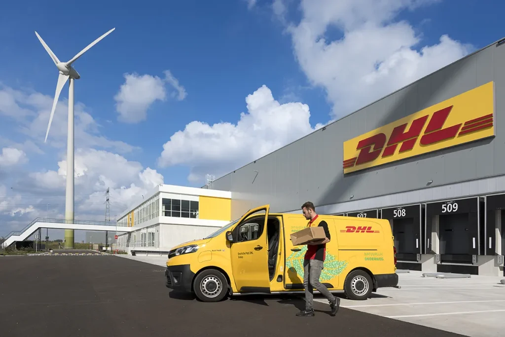 Een DHL-bestelwagen staat geparkeerd buiten een logistieke vestiging van DHL. Een koerier in een grijs uniform met het DHL-logo draagt een pakketje weg van het busje. Het gebouw is modern en wit en het DHL-logo is prominent aanwezig. Op de achtergrond is er een grote windturbine en een gedeeltelijk bewolkte hemel.