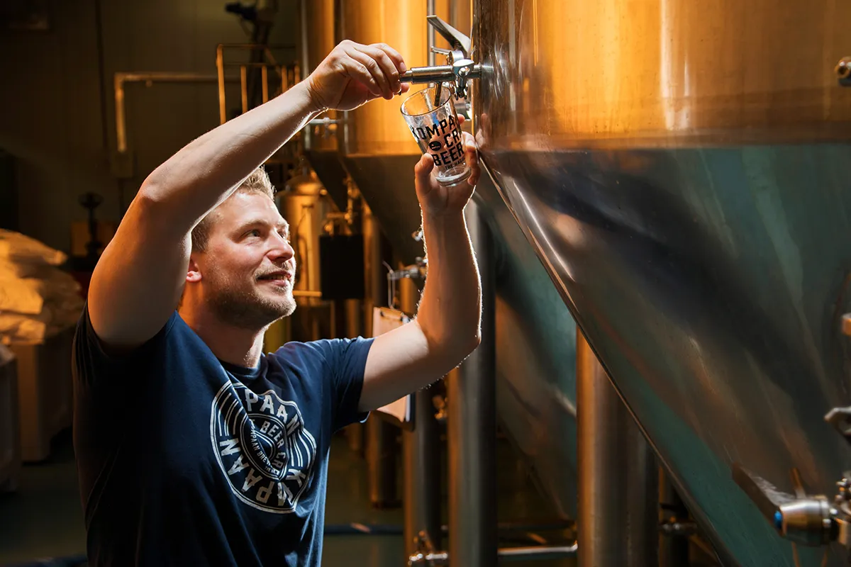 Een man in een blauw shirt vult een glas uit een grote, glimmende metalen brouwtank in een brouwerij. Hij lacht en lijkt gefocust op de taak. De tank heeft leidingen en kleppen, en er zijn nog meer tanks op de achtergrond, wat duidt op een professionele brouwomgeving.