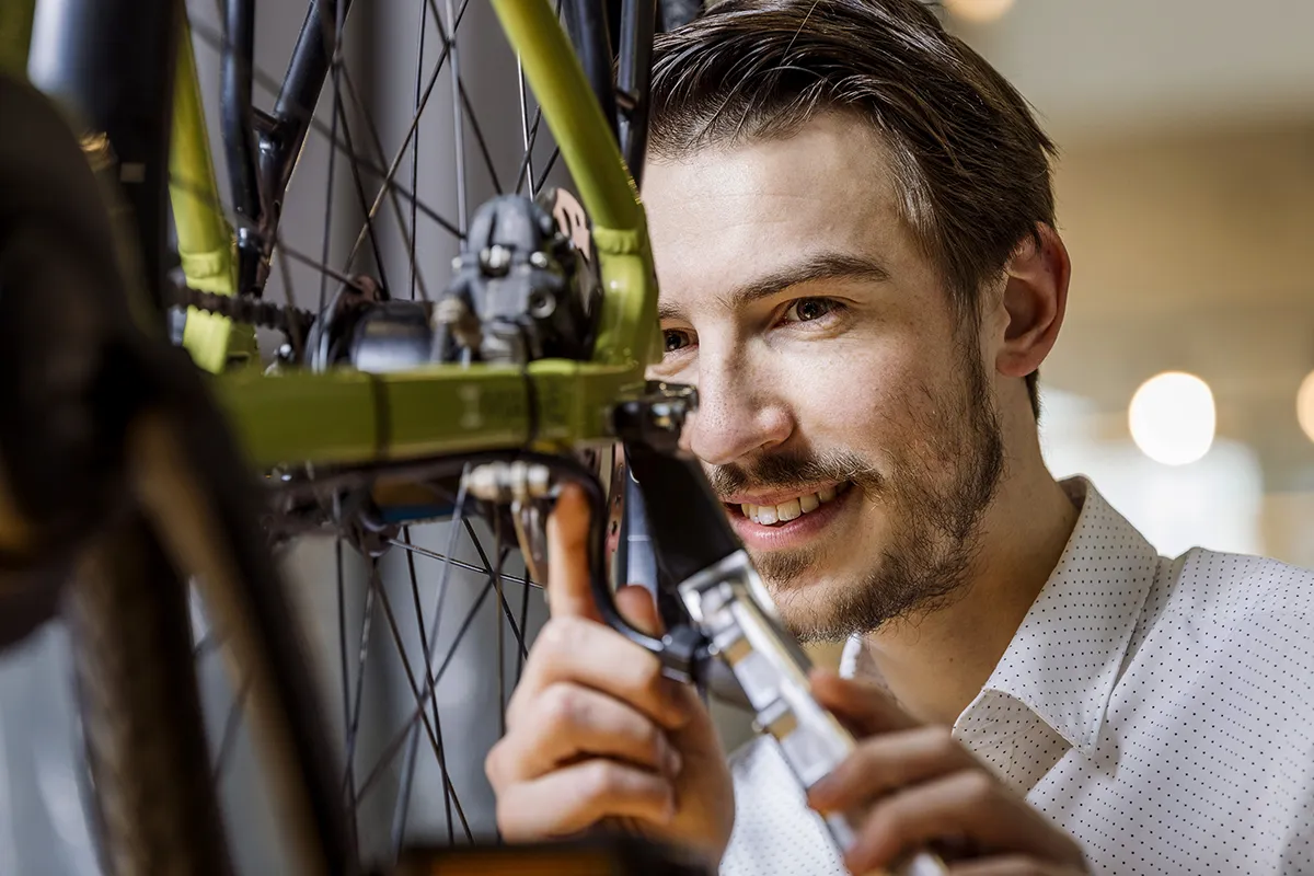 Een persoon met kort bruin haar en een baard glimlacht terwijl hij de versnellingen van een fiets aanpast. Ze gebruiken een stuk gereedschap om aan de fiets te werken en zijn gekleed in een wit shirt met kleine zwarte stippen. De onscherpe achtergrond bevat lichten, wat aangeeft dat de setting waarschijnlijk binnenshuis is.