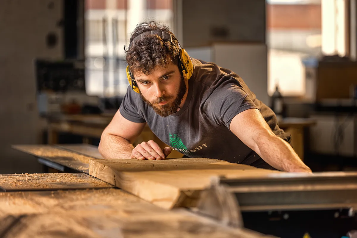 Een man met krullend haar en baard, gele oorbeschermers en een grijs t-shirt draagt, concentreert zich op het schuren van een groot stuk hout. In zijn thuiswerkplaats wordt hij omringd door vage gereedschappen en machines. Hij leunt aandachtig naar voren, volledig verdiept in zijn houtbewerkingsproject.