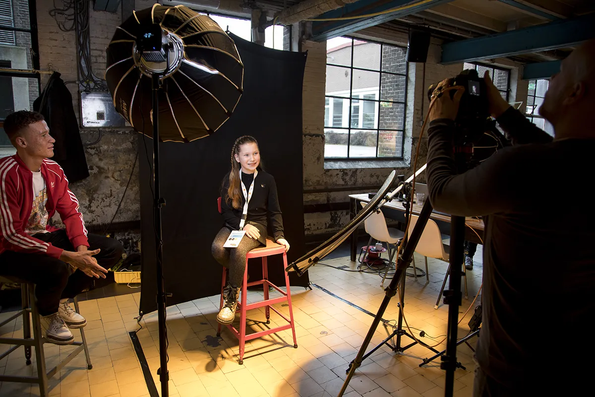 Een jong meisje met lang haar, glimlachend en zittend op een rode kruk voor een zwarte achtergrond in een goed verlichte studio. Een fotograaf maakt een foto van haar terwijl een man in een rood jasje toekijkt. Studioverlichtingsapparatuur is zichtbaar en er staat een tafel met items op de achtergrond.