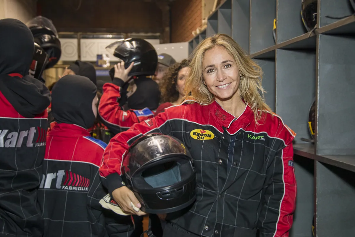 Een glimlachende vrouw met blond haar houdt een zwarte motorhelm vast en staat voor de opbergkasten voor de helm. Ze draagt een zwart en rood racepak met patches, omringd door andere mensen die soortgelijke racepakken en helmen dragen in een indoor kartfaciliteit.