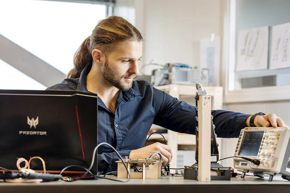 Een man met lang achterover gebonden haar zit aan een bureau in een laboratorium en werkt met een oscilloscoop en elektronische componenten. Verdiept in zijn werk stelt hij met één hand de oscilloscoop in, omringd door diverse elektronische apparaten en een laptop, wat wijst op de nauwgezette aard die vaak in bedrijfsfotografie wordt vastgelegd.