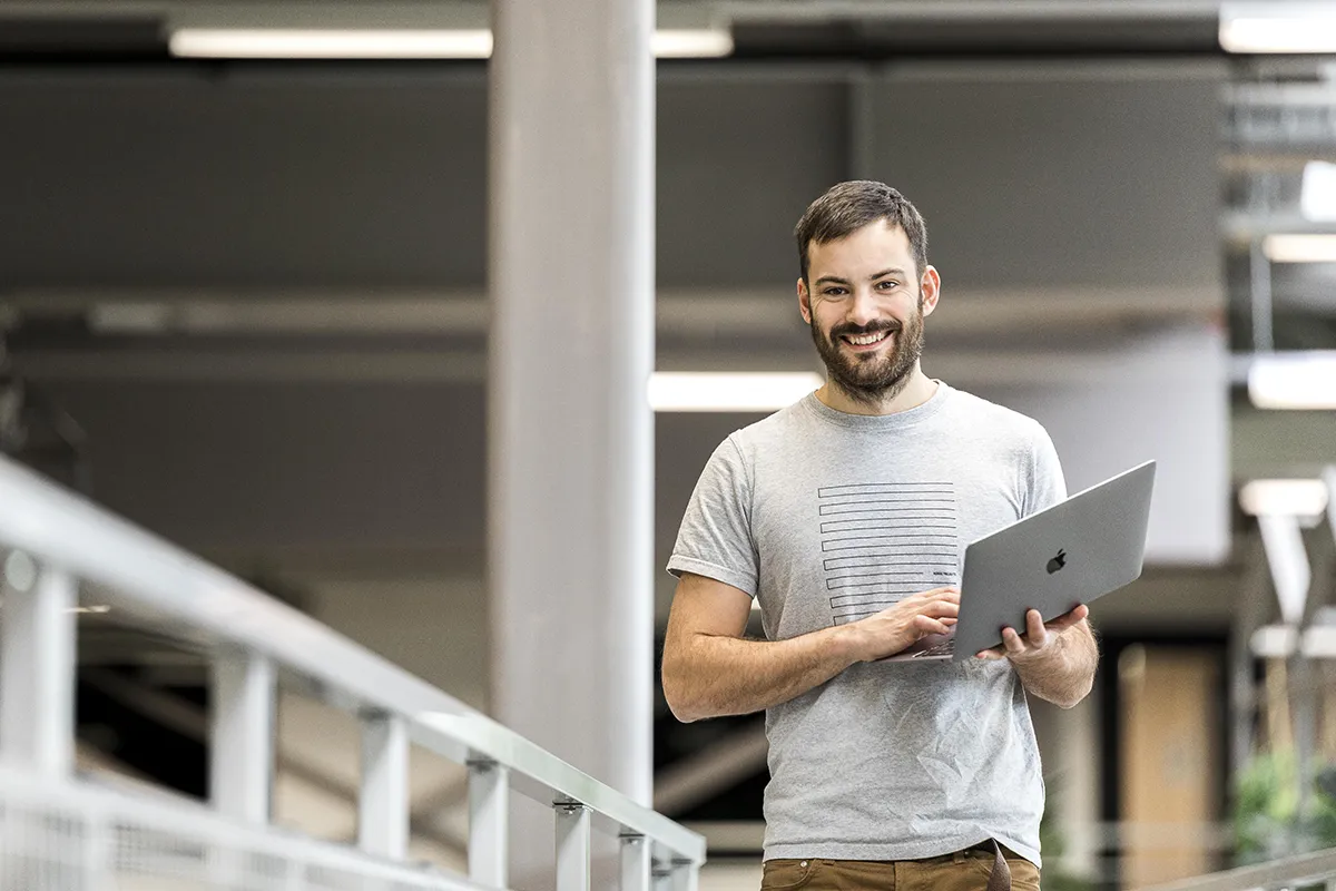 Een bebaarde man in een grijs t-shirt en een bruine broek staat binnen en glimlacht terwijl hij een laptop vasthoudt. Gepositioneerd bij een balustrade bevindt hij zich in een kantooromgeving met moderne designelementen en voldoende verlichting: een uitstekend voorbeeld van bedrijfsfotografie. Het lijkt erop dat hij bezig is met werk of mogelijk met een informeel gesprek.