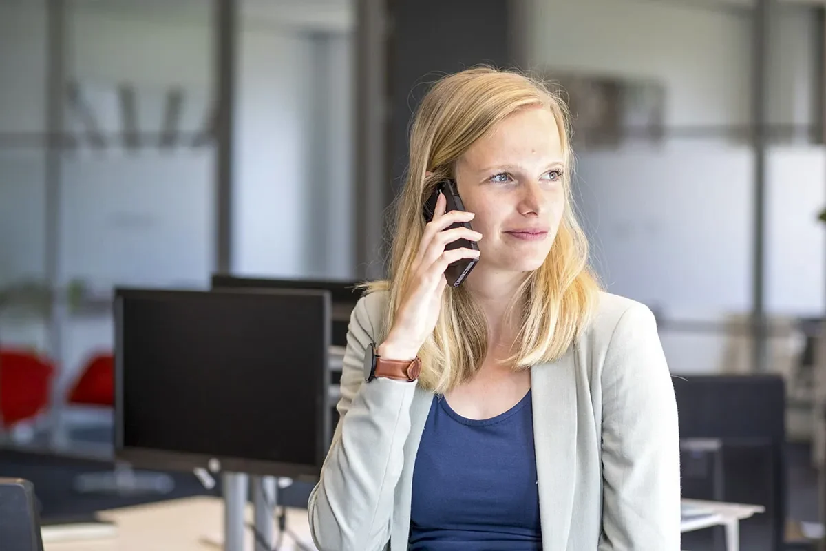 Een vrouw met lang blond haar zit in een kantoor en praat op een smartphone. Ze draagt een lichtgrijze blazer over een blauw topje en een bruin horloge om haar linkerpols. Achter haar staan computerschermen en een glazen scheidingswand, waardoor het kantoor een moderne en professionele uitstraling krijgt: perfect voor bedrijfsfotografie.