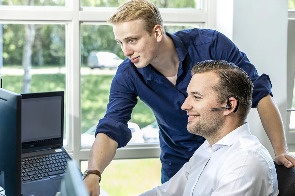 Twee mannen werken samen aan een helder bureau. De man in een blauw shirt staat en wijst naar een laptopscherm, terwijl de zittende man in een wit shirt glimlacht en een koptelefoon draagt. Een groot raam daarachter toont een wazig buitentafereel met bomen en een geparkeerd voertuig, wat de essentie van bedrijfsfotografie weergeeft.