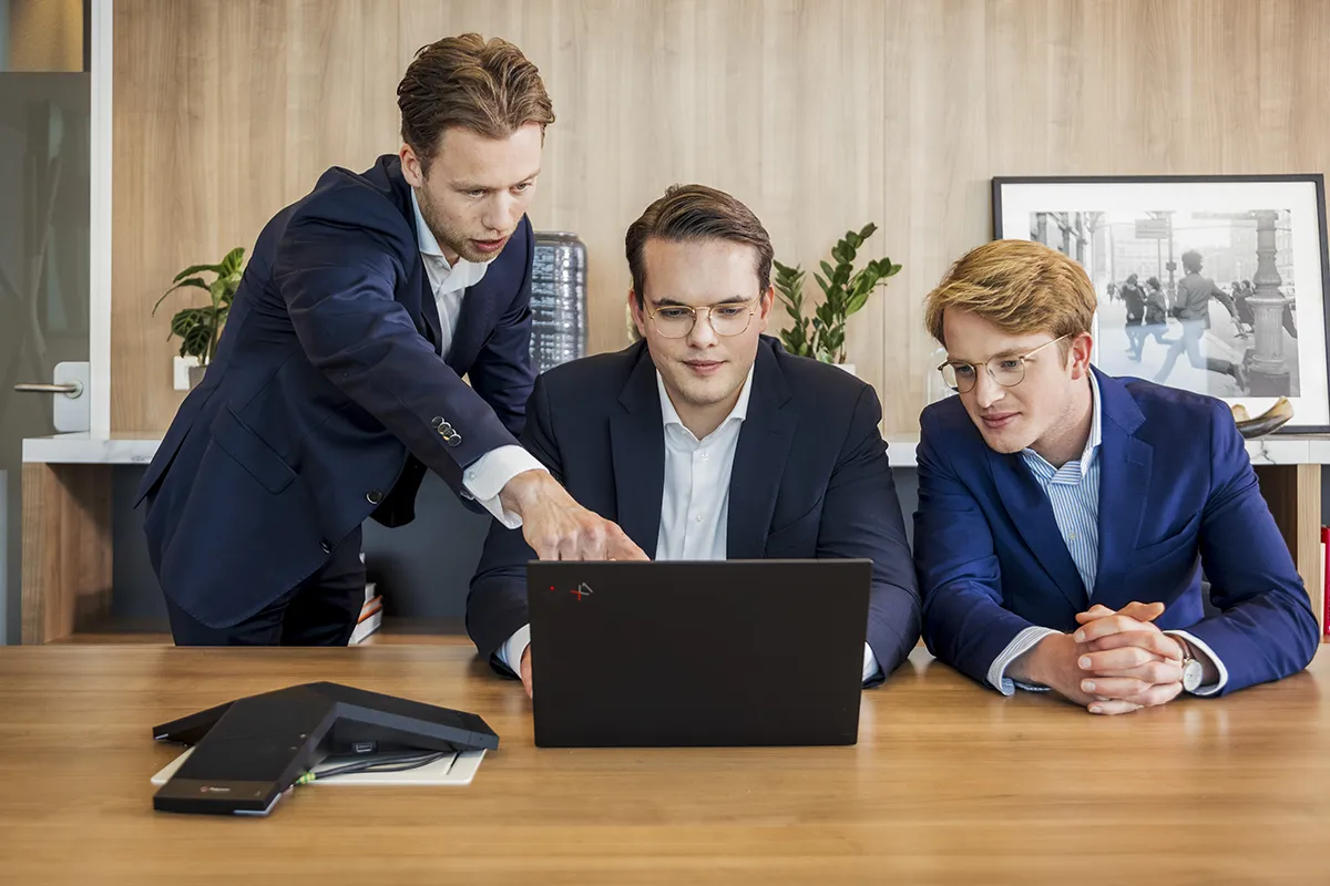 Drie mannen in zakelijke kleding zitten rond een laptop op een houten bureau. De man links staat en wijst naar het scherm, terwijl de andere twee mannen zitten en aandachtig naar de laptop kijken. Een ingelijste foto staat op een plank achter hen, en de achtergrond met houten lambrisering voegt authenticiteit toe aan deze bedrijfsfotografiescène.