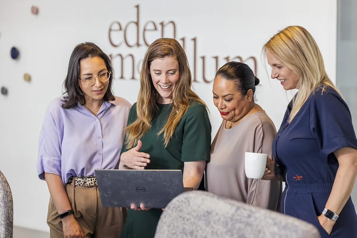 Vier vrouwen staan in een kantoorruimte, verzameld rond een laptop. De vrouw in het midden met lang bruin haar en een groene jurk laat iets zien op het laptopscherm. Ze glimlachen allemaal en zijn betrokken. Op de achtergrond is bewegwijzering te zien met de gedeeltelijk zichtbare tekst "eden memillium." De scène legt perfecte bedrijfsfotografie vast.