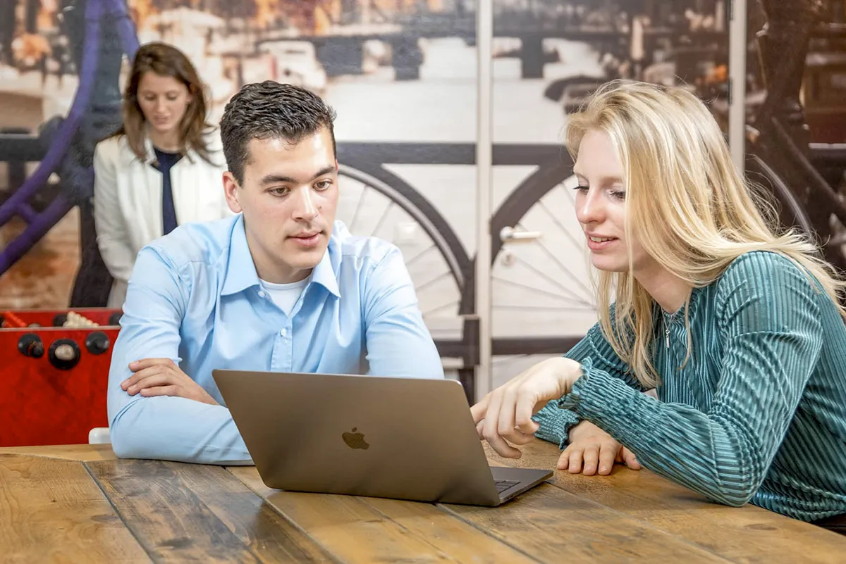 Twee mensen zitten aan een houten tafel en kijken naar een MacBook. De man links, in een blauw shirt, laat zijn armen op de tafel rusten, terwijl de vrouw rechts, in een groene blouse, naar het scherm wijst. In deze bedrijfsfotografiescène staat een vrouw in een witte blazer tegen een muur met op de achtergrond een fietsmuurschildering.