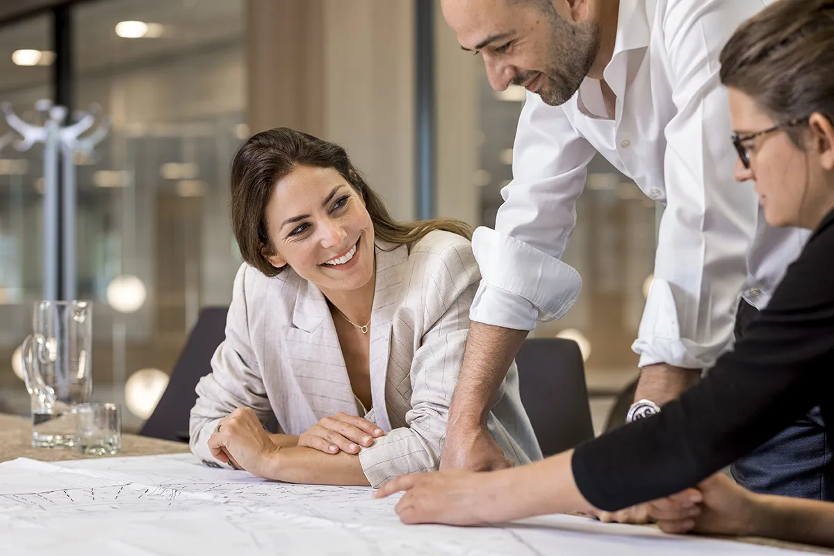 Drie collega's werken rond een tafel samen aan een project. Een vrouw in een lichte blazer lacht naar een man in een wit overhemd die naar een blauwdruk wijst. Een andere persoon, ook met een bril op, bekijkt de blauwdruk. Op de achtergrond zijn een glazen kan en waterglazen zichtbaar: een mooi voorbeeld van bedrijfsfotografie.