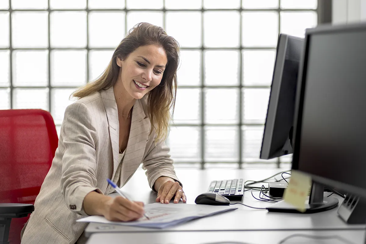 Een vrouw met lang haar, gekleed in een lichtgekleurde blazer, zit aan een bureau met computers. Glimlachend en op een document schrijvend, geniet ze van het natuurlijke licht dat door het grote, betegelde glazen raam naar binnen valt. Het tafereel geeft de essentie van bedrijfsfotografie weer, met een rode stoel gedeeltelijk zichtbaar op de achtergrond.