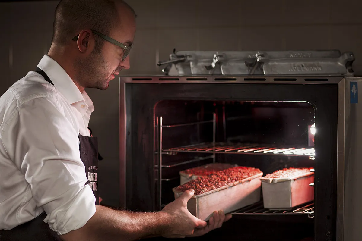 Een persoon met een bril, een wit overhemd en een zwart schort plaatst een pan met rood voedsel in een oven. De ovendeur staat open en er staat al een andere pan met voedsel op het middelste rek. Het lijkt alsof de keuken slecht verlicht is.