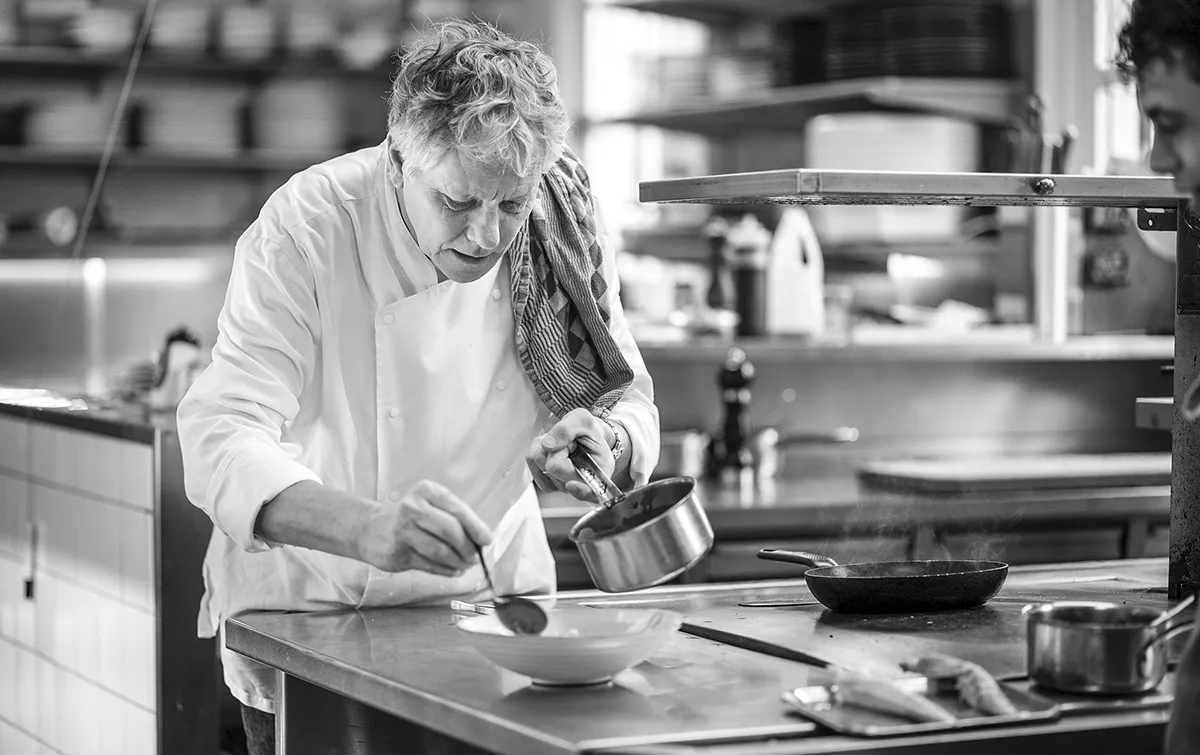 Zwart-wit beeld van een chef-kok die voedsel in een keuken plateert. De chef-kok, gekleed in een witte jas met een handdoek over zijn schouder, giet voorzichtig de saus uit een pan op een schaal. Op het aanrecht zijn een koekenpan en diverse keukengerei zichtbaar. Op de achtergrond zijn planken met keukengerei te zien.