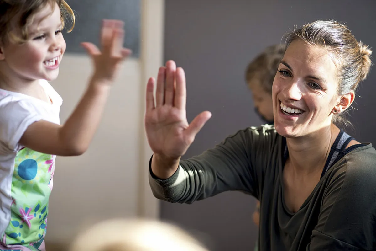 Een glimlachende volwassene met een bruine paardenstaart geeft een high-five aan een vrolijk kind met krullend haar, gekleed in een wit overhemd met kleurrijke patronen. Een ander kind is gedeeltelijk zichtbaar op de achtergrond, wazig en spelend. De achtergrond heeft zachte verlichting en neutrale tinten.