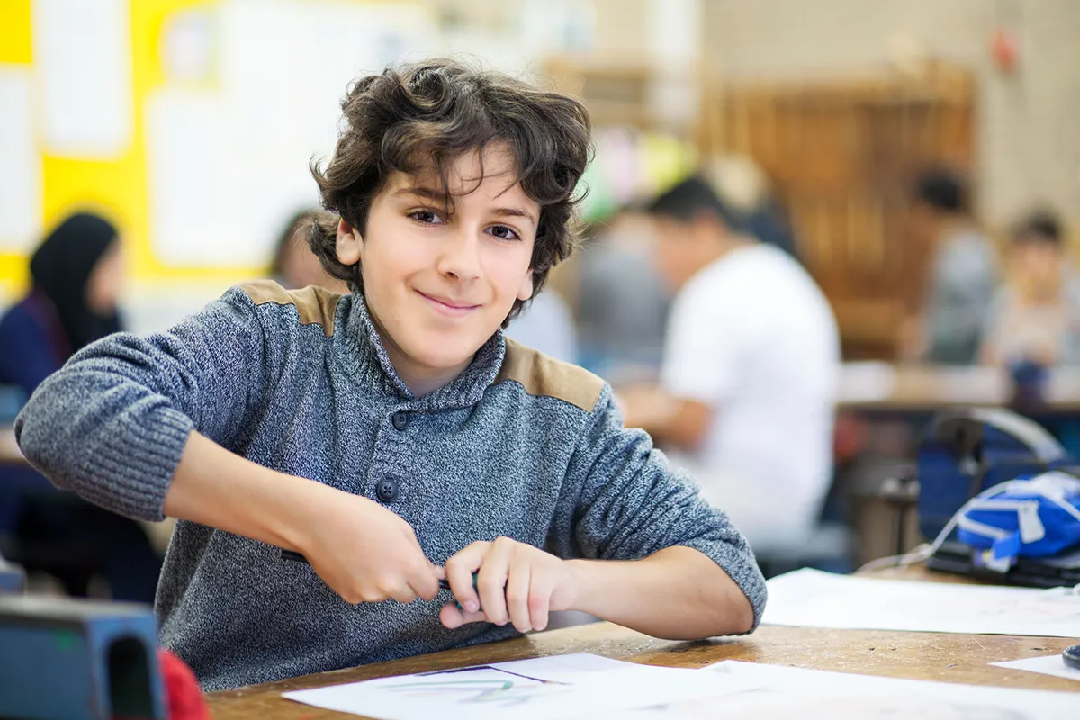 Een jonge jongen met krullend haar, gekleed in een grijze trui, glimlacht terwijl hij aan een klastafel zit. Hij gebruikt een schaar om papier te knippen. Andere studenten en activiteiten in de klas zijn op de achtergrond wazig, wat een drukke, actieve leeromgeving suggereert.