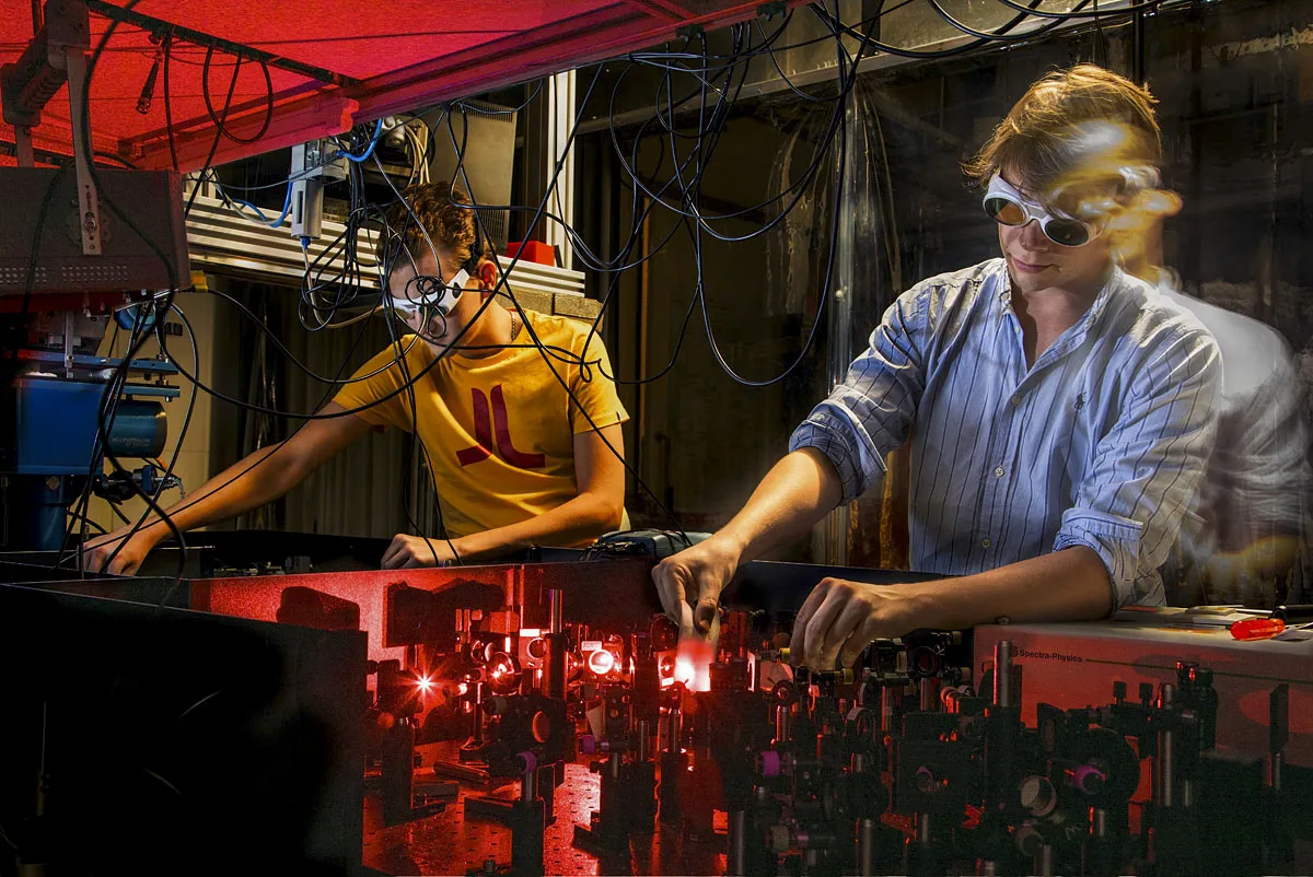 Twee mensen werken in een slecht verlicht laboratorium vol ingewikkelde apparatuur en laserapparatuur. De persoon links draagt een geel shirt en een veiligheidsbril, waarmee hij een onderdeel afstelt. De persoon rechts, in gestreept shirt en veiligheidsbril, hanteert een ander deel van de opstelling, verlicht door rood laserlicht.